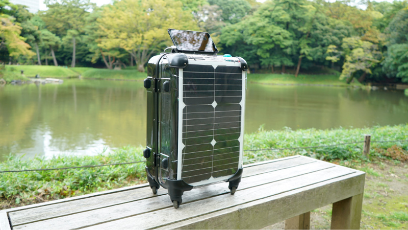 A medium-sized luggage with solar panels on the sides stands on a bench in front of a lake at a park.