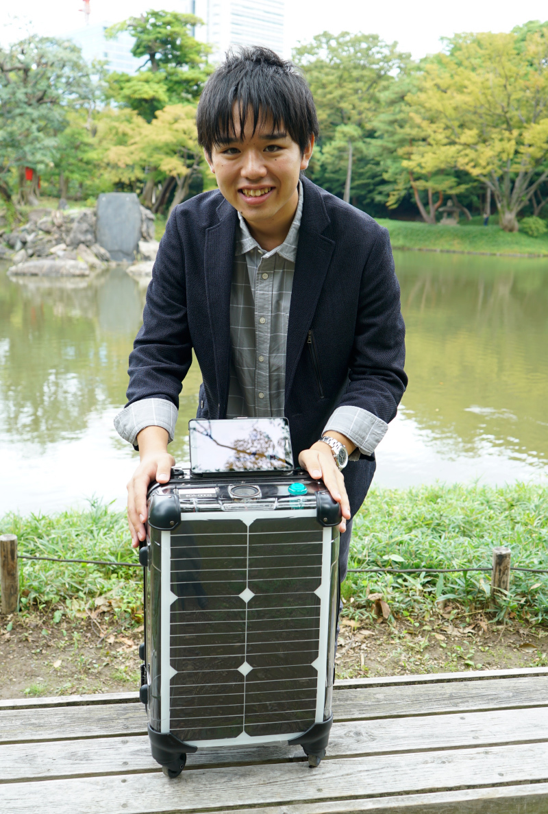 Kazumi Muraki stands with a smile behind a medium-sized luggage with solar panels on the sides stands on a bench in front of a lake at a park.