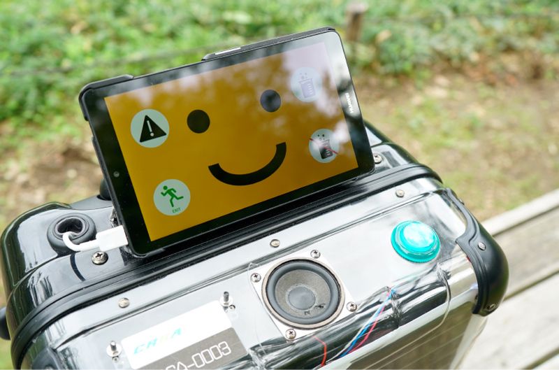 A medium-sized luggage with solar panels on the sides and a tablet with a orange smiley face on top stands on a bench in a park.