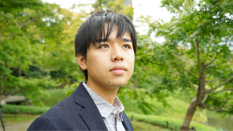 Young Kazumi Muraki looks up to the sky wearing a suit in a green lush park.