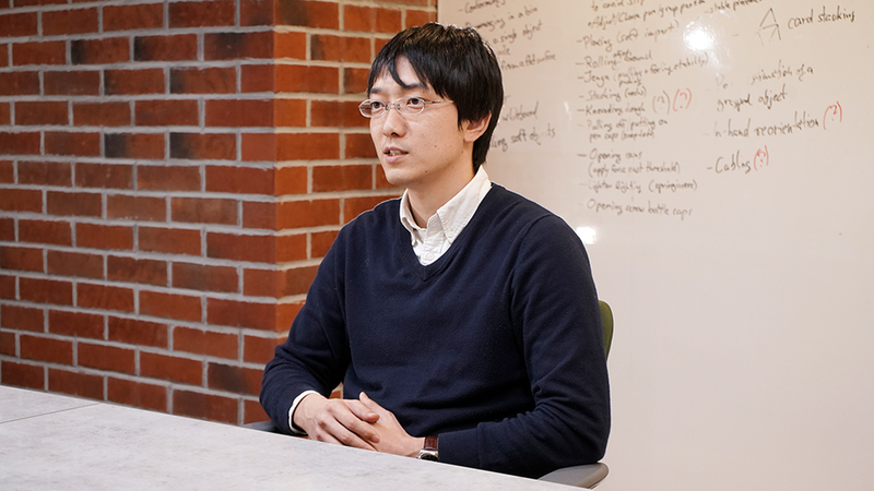 Kazutoshi Tanaka sits at a desk in front of a whiteboard with his hands interlocked. He is wearing glasses and a black shirt.