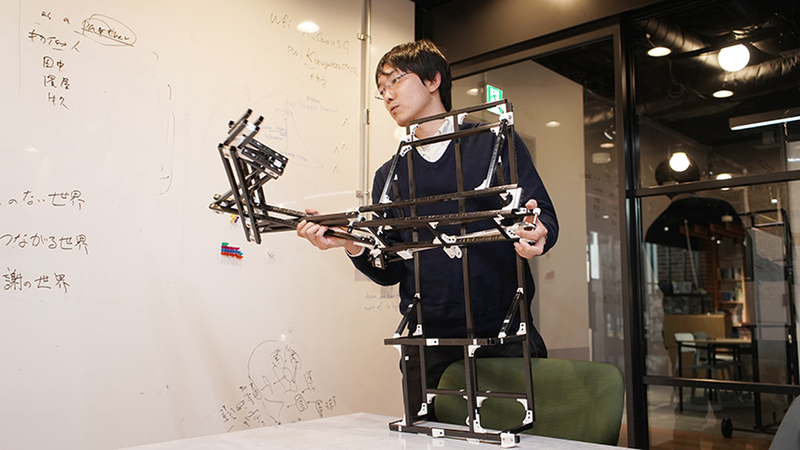 Kazutoshi Tanaka grabs a robotic frame made of thin black metal bars. 