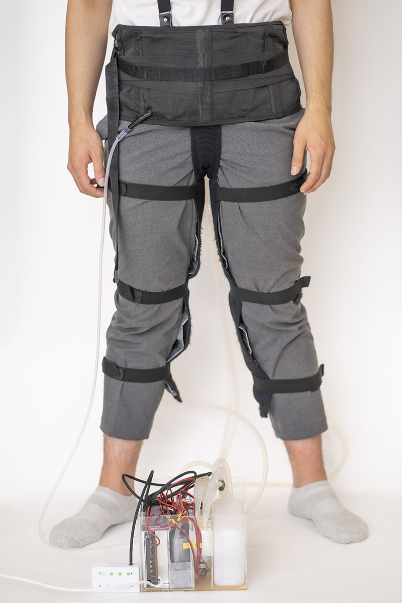 A man stands in a white room wearing a harness that is connected to a device with tubes