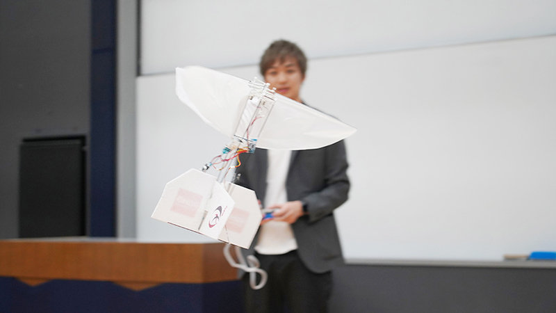 A white lightweight robot flaps it's wings to hover infront of a man in a classroom of a university