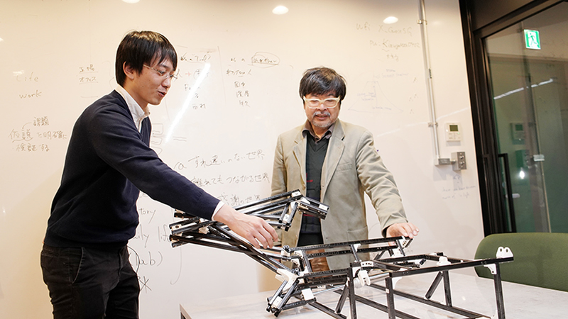 Kazutoshi Tanaka demonstrates the robotic frame's movement to the interviewer, Satoshi Endo