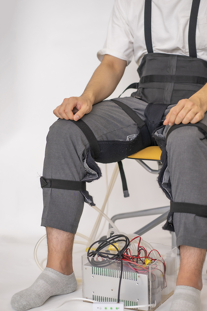 A man sits in a chair in a white room wearing a harness that is connected to a device with tubes