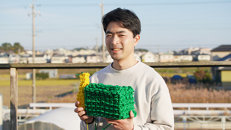 Hirose Yuichi standing outside holding two of his solid knitting creations.