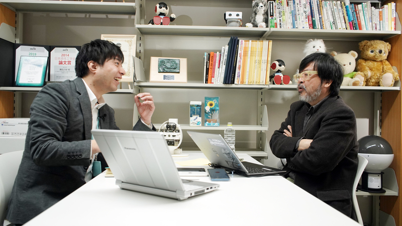 Yoshihiro Sejima sits at a desk across from an interviewer, Satoshi Endo. A bookshelf filled with books and small robots is in the background.