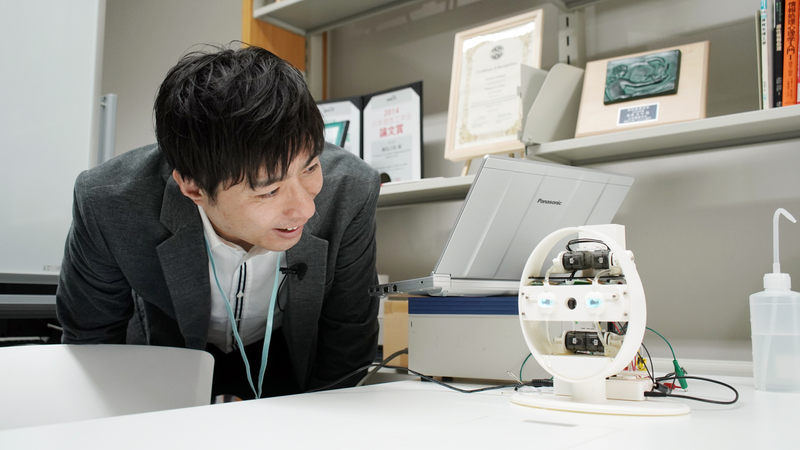 Yoshihiro Sejima leans over and looks at a small circular robotic device with human-like eyes.