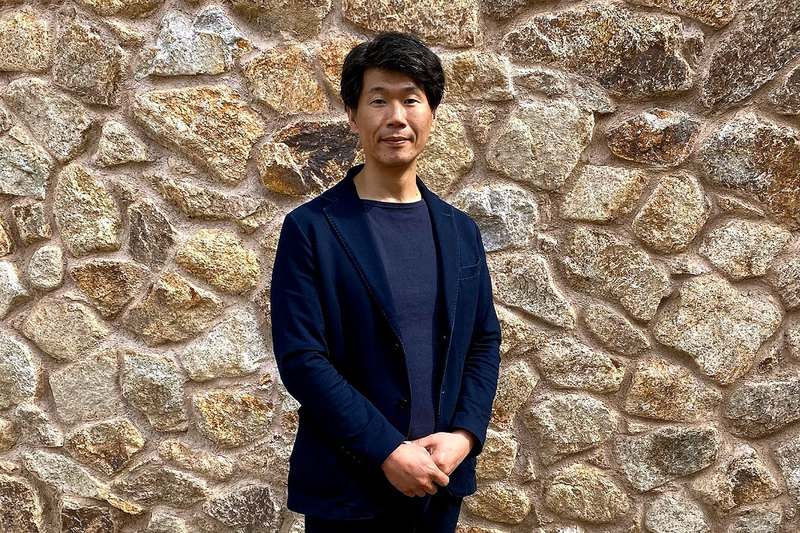 Yusuke Nakano stands in front of a brick wall, he is wearing smart casual clothes